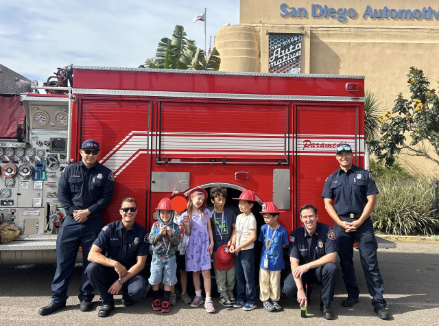 Kids during the Summer Camp standing with firefighters and their fire truck, in front of the San Diego Automotive Museum building.